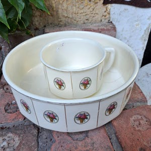 Op de afbeelding: Crème emaille wastafel en kan set. De ronde wastafel heeft bloemenmand decals. De kan heeft een handvat en bijpassende bloemen decals. De set is vintage en vertoont gebruikssporen.