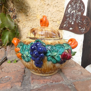 Può includere: Una zuppiera in ceramica con coperchio, con un design di frutta. La zuppiera è beige con dettagli marroni e manici arancioni. Uva blu, ciliegie rosse e foglie verdi adornano i lati. Il coperchio ha una maniglia arancione.