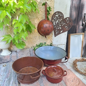 Op de afbeelding: Een verzameling vintage keukengerei in bruine en rode tinten. Inclusief een vergiet, een grote kom met een witte binnenkant, een kleine pot met deksel en een metalen bak met een geperforeerde bodem. Een decoratieve hartvormige metalen wandhanger is ook zichtbaar.