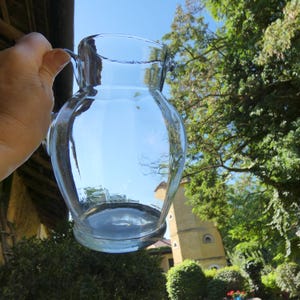 Pode incluir: Uma jarra de vidro transparente com corpo arredondado, boca larga e uma alça resistente na lateral. O recipiente apresenta uma superfície lisa e translúcida que reflete a luz ambiente, exibindo um design simples e funcional.