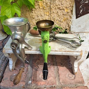 Pode incluir: Duas ferramentas de cozinha antigas são exibidas em uma pequena mesa de madeira branca desgastada. Uma é um moedor de carne prateado com uma alça de madeira. A outra é um moedor de alimentos verde brilhante com uma tigela de latão e uma alça preta. Um batedor de metal também é visível.