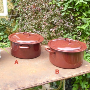 Puede incluir: Dos ollas de esmalte rojo con tapas, etiquetadas como A y B, sobre una mesa de metal oxidada.