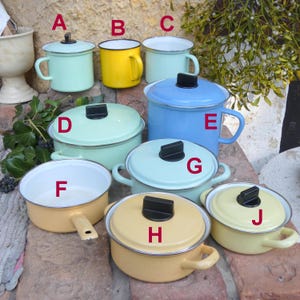 Könnte beinhalten: Eine Sammlung farbenfroher Emaille-Töpfe und -Becher. Das Sortiment umfasst hellblaue, gelbe und blaue Teile mit schwarzen Griffen und Deckeln. Einige haben Griffe, andere seitliche Griffe. Die Artikel sind auf einer Ziegeloberfläche angeordnet.