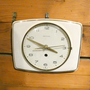 Puede incluir: Un reloj de pared rectangular blanco con ribete y manecillas doradas. La esfera del reloj tiene números negros y la marca "Hettich". El reloj está montado en una pared de madera con soportes metálicos.