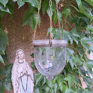Könnte beinhalten: Eine transparente Glashängelaterne mit geätzten Blumen- und Traubenmotiven, die an einer Metallkette aufgehängt ist. Die Laterne hat einen abgerundeten Boden und einen Deckel. Der Hintergrund zeigt grünes Efeu und eine Statue der Jungfrau Maria.