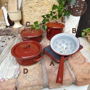Könnte beinhalten: Eine Sammlung von Vintage-Küchenutensilien, darunter eine rote Emaille-Colander, ein kleiner Topf mit Deckel und ein Henkelbecher. Die Colander hat einen weißen Innenraum mit einem Muster aus kleinen Kreisen. Die Artikel sind auf einer Ziegeloberfläche angeordnet.