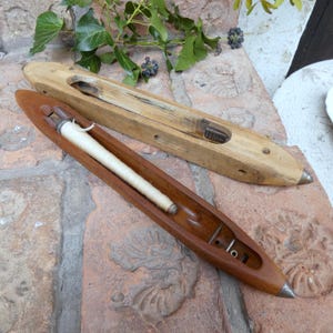 Peut inclure: Deux navettes de tissage en bois. L'une est en bois clair, l'autre en bois plus foncé avec une bobine de fil blanc. Les deux ont des embouts métalliques et sont conçues pour le tissage de textiles. Les navettes sont posées sur une surface en briques.