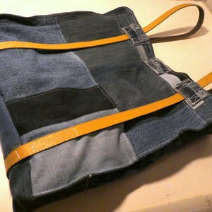 Op de afbeelding: Een patchwork tote bag gemaakt van denim stof met gele leren banden. De tas is gemaakt van verschillende tinten blauw denim en heeft een patchwork ontwerp.