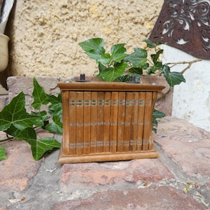 May include: A vintage wooden box with a series of small, vertical compartments, possibly for storing small items. The box is made of light brown wood and has a decorative design on the front. The box is sitting on a brick surface.