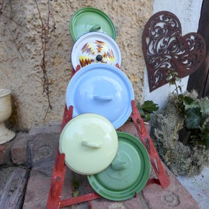 Pode incluir: Cinco tampas de panela de esmalte vintage em várias cores e padrões, incluindo verde, azul, amarelo e um design geométrico, são exibidas em um suporte de metal vermelho.