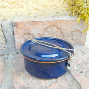 Può includere: Un contenitore rotondo in smalto blu navy con coperchio e manico in metallo. Il contenitore ha un bordo arancione e una chiusura in metallo argentato. Il contenitore è su una superficie di mattoni.