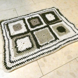 Op de afbeelding: Een handgemaakt gehaakt vloerkleed met een geometrisch patroon van vierkanten in wit, zwart en grijs. Het vloerkleed heeft een textuur en een rand die het vierkante ontwerp aanvult. Dit decoratieve vloerkleed is geschikt voor diverse ruimtes.