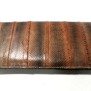 Può includere: Una pochette in pelle di serpente marrone con un motivo testurizzato. La pochette è chiusa e ha una cucitura cucita attorno ai bordi.