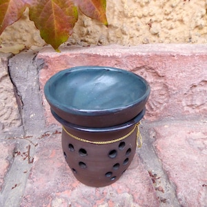Könnte beinhalten: Ein handgefertigter Keramik-Duftlampenhalter mit einer dunkelblauen Schale und einem braunen Sockel. Der Sockel hat dekorative Löcher und ist mit einer goldfarbenen Schnur verziert. Der Halter steht vor einem Hintergrund aus Stein und Laub.