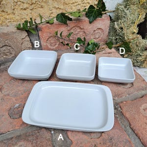 Peut inclure: Ensemble de quatre plats en céramique blanche. Les plats sont de forme carrée et rectangulaire. Les plats sont disposés en rangée sur une surface en brique.
