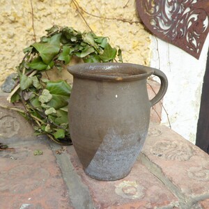 Peut inclure: Un pot en terre cuite brun avec une anse. Le pot a une surface rugueuse et texturée et est légèrement décoloré.