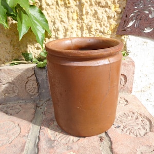 Op de afbeelding: Een bruine, cilindervormige keramische pot met een licht uitlopende rand. De pot heeft een glad oppervlak en een scheur aan de zijkant. Hij staat op een bakstenen oppervlak, met groene bladeren op de achtergrond. De pot is waarschijnlijk voor opslag of decoratie.