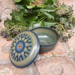 Puede incluir: Un cuenco de cerámica con su tapa a juego. La tapa presenta un diseño floral azul, naranja y verde. El cuenco y la tapa son de tonos verdes y marrones. El cuenco está abierto, revelando el interior. Hiedra y bayas en el fondo.