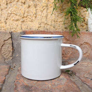 Może przedstawiać: Biały emaliowany kubek z niebieską obwódką i uchwytem. Kubek ma klasyczny design z solidnym uchwytem, idealny do kawy lub herbaty. Kubek jest ustawiony na tle cegieł.