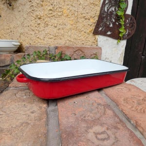 May include: A rectangular enamel roasting pan with a bright red exterior and a smooth white interior. The pan features a thin black rim along the top edge and two small, curved red handles on the sides for gripping. The surface is glossy and solid.