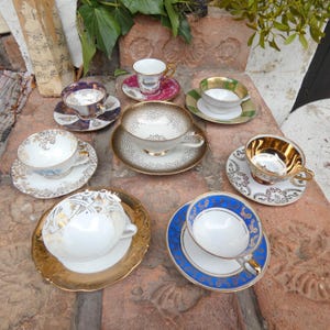 Può includere: Una collezione di tazze da tè e piattini vintage in vari design. Le tazze presentano bordi dorati, motivi floreali e accenti colorati. I piattini sono bianchi, dorati e blu. Le tazze da tè sono disposte su una superficie di mattoni.