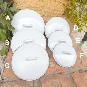 Op de afbeelding: Een verzameling van zes witte emaille pannendeksels in verschillende maten. Elke deksel heeft een ronde vorm met een kleine, gebogen handgreep. De deksels zijn gerangschikt op een bakstenen oppervlak, met groen in de achtergrond.