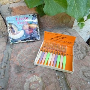 Può includere: Un set di dodici forchettine in acciaio inossidabile con manici in plastica colorata nelle tonalità crema, viola, rosa e verde, disposte in una scatola arancione con l'etichetta "STAINLESS STEEL LITTLE FORKS" accanto a una scatola vintage.