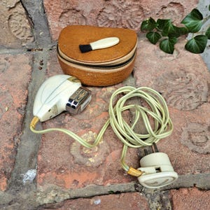 Könnte beinhalten: Ein Vintage-Elektro-Rasierer mit einem weißen Kunststoffgehäuse und einer silbernen Metallklinge. Der Rasierer ist an ein Kabel mit einem weißen Stecker angeschlossen. Der Rasierer befindet sich in einem braunen Lederetui mit einer kleinen Bürste.