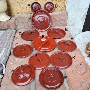 Könnte beinhalten: Eine Sammlung von 13 roten Emaille-Deckeln mit verschiedenen Designs, darunter ein Deckel mit einem Sieb, ein Deckel mit einem Griff und ein Deckel mit einem geteilten Abschnitt. Die Deckel sind auf einer Ziegelstein-Oberfläche angeordnet.