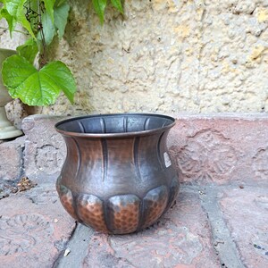 Puede incluir: Una maceta decorativa de color cobre, con un borde acanalado y una base bulbosa. La parte inferior de la maceta tiene un aspecto texturizado y martillado. Descansa sobre una superficie de ladrillo, con hojas verdes y una pared de piedra en el fondo.