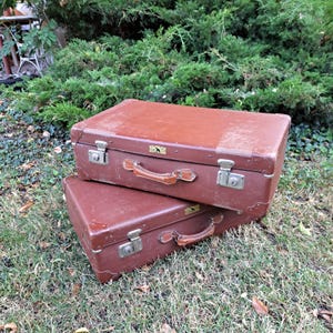 Op de afbeelding: Twee vintage bruine koffers op elkaar gestapeld. De koffers hebben metalen sluitingen, leren handvatten en een klein decoratief embleem. Ze staan op gras, omringd door groen. Gebruikssporen.