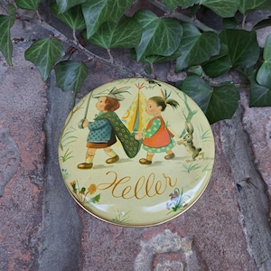 Può includere: Una scatola rotonda di colore crema con un'illustrazione colorata di due bambini che camminano con un coniglio. La scatola ha la parola "Heller" scritta in corsivo sul davanti.