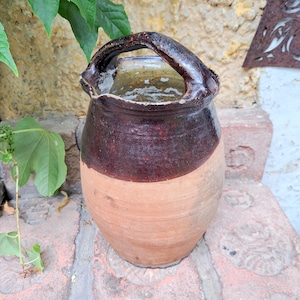 May include: A rustic pottery jug with a dark brown upper section and a light brown lower section. It features a large, arched handle and a wide opening. The unglazed pottery has a textured surface, possibly an antique or vintage piece. The jug is likely for decorative use.