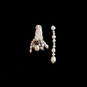 Puede incluir: Un par de pendientes elegantes con perlas blancas brillantes y una gema de color púrpura intenso. Un pendiente tiene un grupo de perlas con una gema central, el otro es una larga hebra de perlas. Sobre un fondo negro.