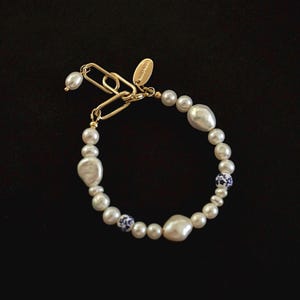 Puede incluir: Un brazalete de cadena dorada con cuentas de perlas blancas y cuentas con patrones azules y blancos.
