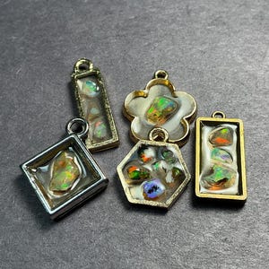 May include: A collection of four metal pendants featuring iridescent opal fragments embedded in a white resin base. The set includes a silver-toned diamond shape, a gold-toned flower shape, a gold-toned hexagon, and a gold-toned rectangular pendant.