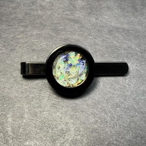 May include: A black tie clip with a circular centerpiece. The centerpiece features a design of iridescent, colorful fragments set in a black frame. The clip is made of black metal and is set against a gray background.