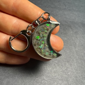 May include: A crescent moon-shaped keychain with a silver-colored metal frame. The moon is filled with a mix of dark and light-colored fragments, with hints of green and blue. The keychain has a clasp and a ring for attaching keys.