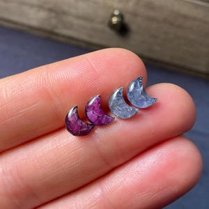 May include: Four pairs of crescent moon-shaped earrings in shades of purple and blue. The earrings are small, with a polished, gemstone-like appearance. The earrings are displayed on a person's fingers, with a blurred background.