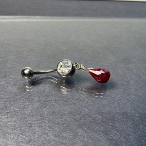 May include: A curved belly button ring with a silver-colored barbell. One end features a clear, round gemstone, and the other has a dangling, faceted, teardrop-shaped, deep red gemstone. The jewelry is set against a gray background.