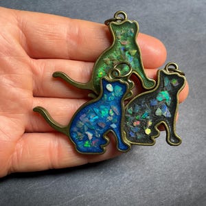 May include: Three cat-shaped pendants with a bronze-colored outline. The cats are filled with colorful, iridescent material. One cat is green, one is blue, and one is black. Each pendant has a small loop at the top for hanging.