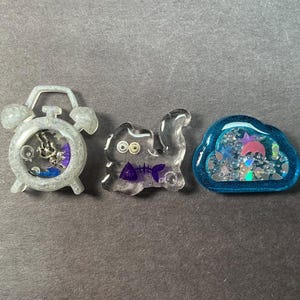 May include: Three resin shaker charms: a gray alarm clock, a clear cat, and a blue cloud. The alarm clock has a silver clock face with purple and blue accents. The cat has purple accents. The cloud has a pink umbrella.