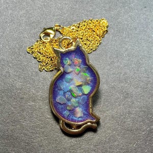 May include: A gold-colored cat-shaped pendant necklace with a blue background filled with colorful, iridescent stones. The pendant is attached to a delicate gold chain. The necklace is displayed on a dark gray surface.