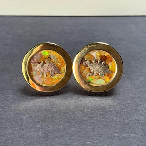 May include: A pair of gold-toned cufflinks. Each cufflink features a round design with a dinosaur silhouette in the center. The background has a mix of orange, yellow, and green tones. The cufflinks are set against a dark background.