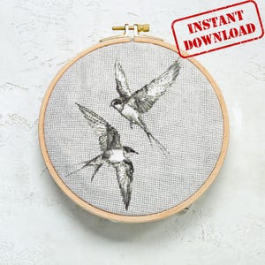 Puede incluir: Un aro de bordado de punto de cruz con un diseño de dos pájaros negros y blancos en vuelo. Los pájaros son estilizados y tienen alas largas y puntiagudas.