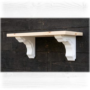 May include: A wall-mounted shelf featuring a natural light-coloured wood plank top supported by two white decorative corbels with curved, scalloped edges. The shelf is positioned horizontally, showcasing the wood grain on the top surface.