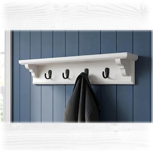 May include: A white wall-mounted shelf featuring four matte black metal hooks. The shelf has decorative side brackets and a flat top surface. A dark grey garment hangs from one of the hooks, partially obscuring the centre of the unit.