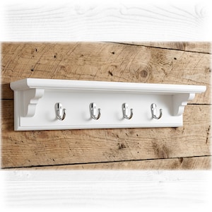 May include: A white wall-mounted shelf featuring a flat top surface and four polished silver metal hooks attached to the front panel. The shelf has decorative side brackets and a rectangular backboard, providing a functional storage solution.