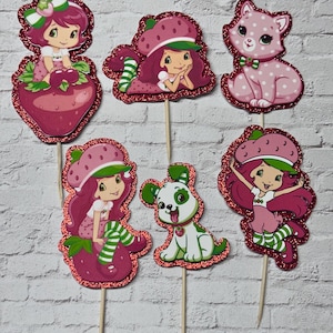 May include: A collection of six glittery, die-cut toppers featuring Strawberry Shortcake characters. The toppers include Strawberry Shortcake, a cat, and a dog, all in shades of pink, red, green, and white, mounted on wooden sticks for easy insertion.