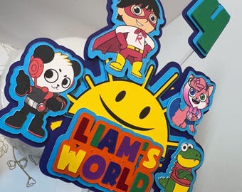 May include: A colorful cake topper featuring a cartoon sun with a smiling face, the words "Liam's World", a panda wearing headphones, a red superhero, a pink cat, and a green alligator.